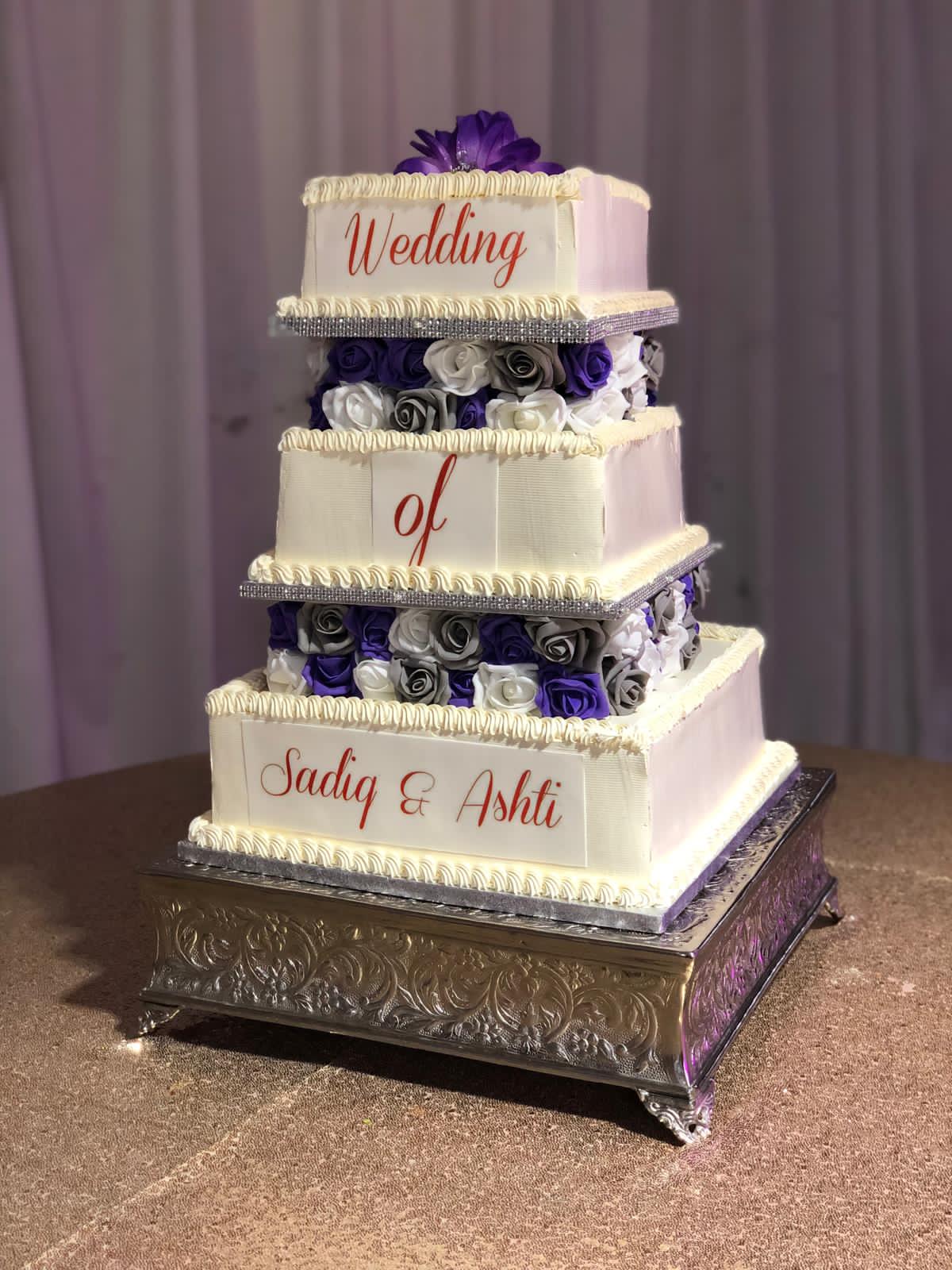Four-tiered wedding cake with purple and white flowers on a metallic stand.