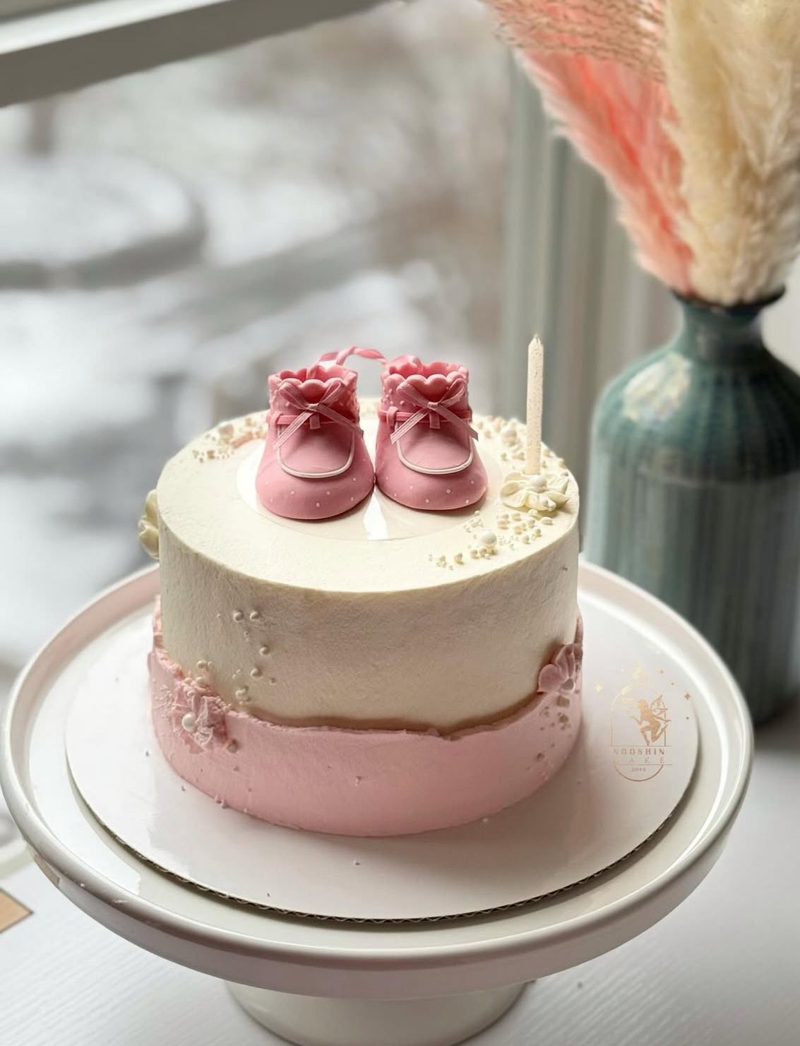 Small pink and white cake with baby shoes and a candle on a white surface.