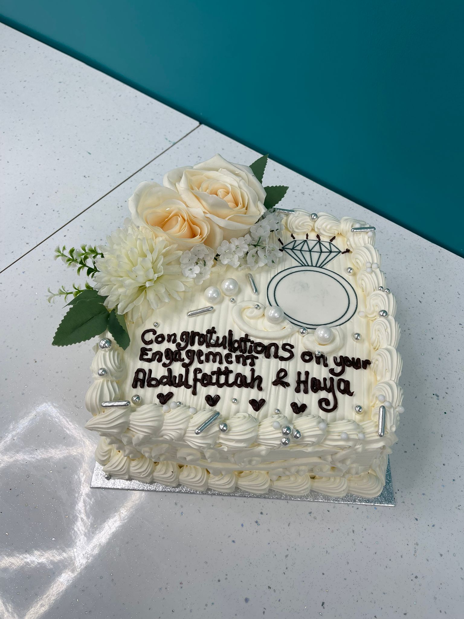 Engagement cake with white frosting, flowers, and a message on a light surface.