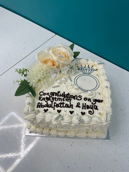 Engagement cake with white frosting, flowers, and a message on a light surface.