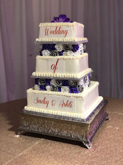 Four-tiered wedding cake with purple and white flowers on a metallic stand.