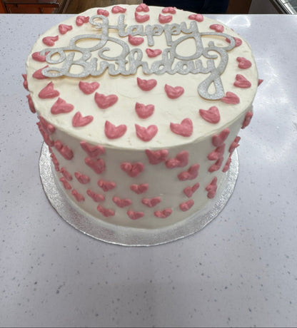 White birthday cake with pink hearts and 'Happy Birthday' text on a white surface.